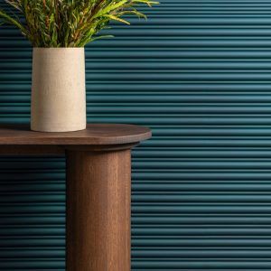 A teal 3D wall panel with horizontal ribbed detailing. In the foreground, a dark wooden table with rounded edges holds a beige vase containing green leafy plants.